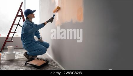 female painter in overalls with cap and gloves painting a wall with paint roller, ladder and buckets on background. Copy space Stock Photo