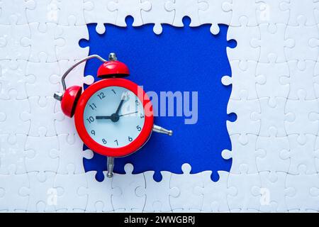 Red alarm clock sitting on a white jigsaw puzzle with a blue missing piece area Stock Photo