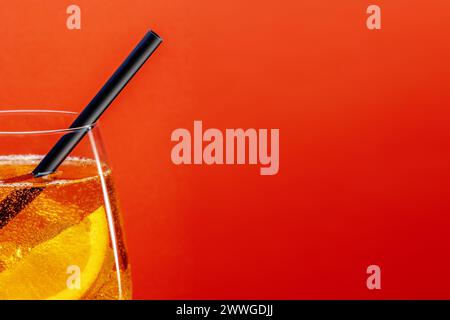 Detail of aperol spritz aperitif with orange slice, ice cubes and black drinking straw on red background with space for text. Stock Photo