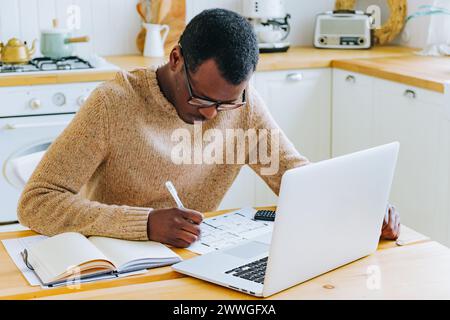 A man is deeply engrossed in planning, pen in hand, with a laptop and notebook at his kitchen table. His focus is indicative of careful thought and organization. The designer is developing a new apartment renovation project. Stock Photo