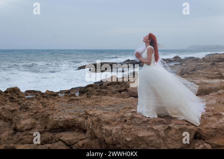 Lonely woman in wedding dress praying to gods a groom for wedding on windy day on Mediterranean sea coast, Alicante, Spain Stock Photo