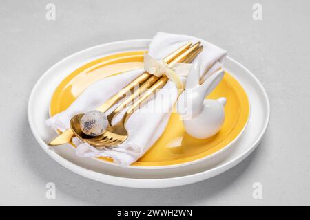 Yellow and white plates, white napkin with gold cutlery and quail egg, white ceramic bunny on them. Neutral light gray background. Idea of serving tab Stock Photo
