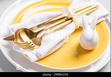 Golden cutlery in white napkin and white ceramic bunny on yellow white plates. Easter table setting concept. Passover table serving. Close up. Horizon Stock Photo