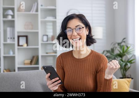 Cheerful young woman with eyewear holding smartphone and making a triumphant gesture in a cozy living room setting. Stock Photo