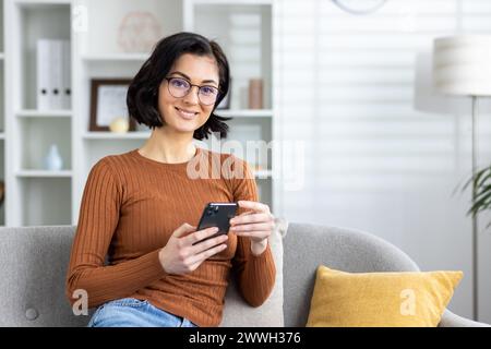 Young woman in casual wear engaged with her smartphone, sitting comfortably in a bright, modern living room setting. Stock Photo