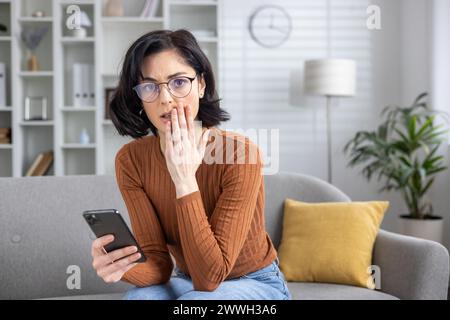 Worried female sitting on a couch, looking confused at her phone after receiving unsettling news. Stock Photo