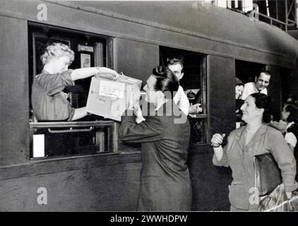 young man smoking cigarette hands cardboard box labeled „baby“ through train window to blonde woman inside the train , railway station 1961 Germany Stock Photo