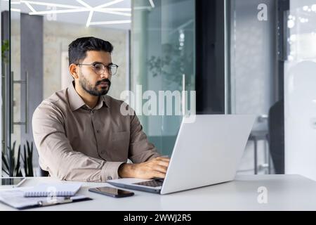 Focused Indian man in smart casual attire working and thinking seriously while using a laptop in a modern office setup. Stock Photo