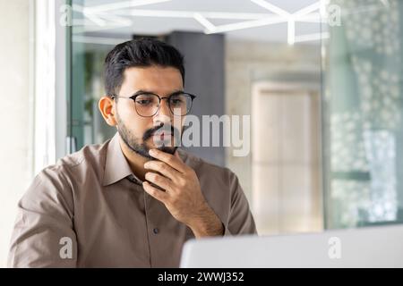 Young Indian businessman in a serious thinking pose, working on his laptop at an office, showing dedication and focus. Stock Photo