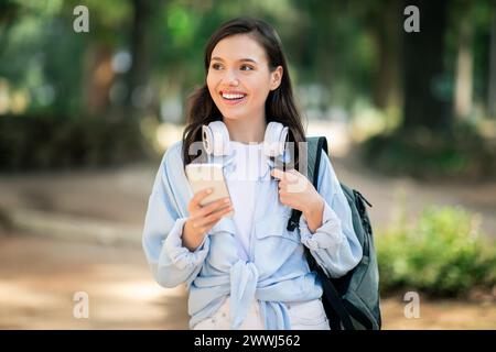 Smiling young student with headphones around her neck using a smartphone in a sun-drenched park Stock Photo