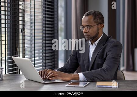 Serious focused male businessman working inside office with laptop, mature experienced african american man typing on keyboard, thinking reading from monitor. Stock Photo