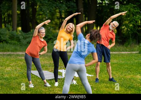 Group of seniors following yoga stretch routine outdoors, led by younger instructor Stock Photo