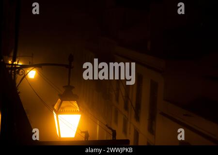 Mysterious Foggy Night in Old Town with Vintage Street Lamp. A warm glow emanates from an antique street lamp on a foggy night, the mist veiling the outlines of buildings behind. Stock Photo