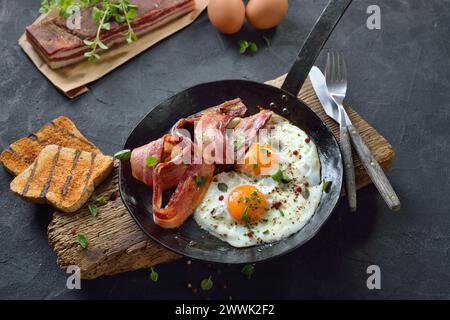 Crunchy fried eggs with Italian pancetta bacon served  in an old iron frying pan on rustic weathered wood Stock Photo