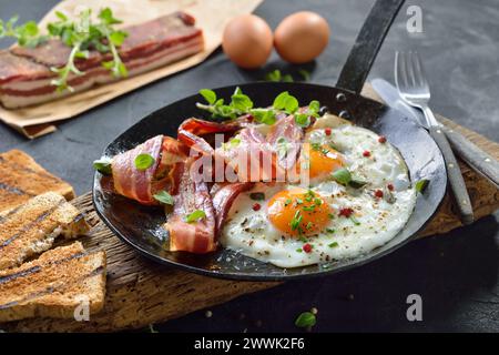 Crunchy fried eggs with Italian pancetta bacon served  in an old iron frying pan on rustic weathered wood Stock Photo