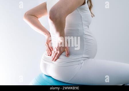 Description: Pregnant woman with baby bump feels back pain and holds hands on her aching back while sitting on gym ball. Final month of pregnancy - we Stock Photo