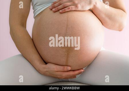 Description: Mother sits on a stool and gently holds her very round pregnant baby bump. Front view. Pink background. Bright shot. Stock Photo