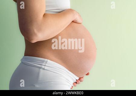 Description: Midsection of unrecognizable standing woman gently holding her very round pregnant baby bump. Side view. Green background. Bright shot. Stock Photo