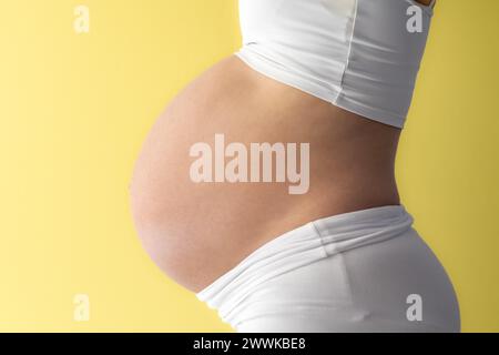 Description: Midsection of unrecognizable standing mother in white cloths with very round pregnant baby belly. Side view. Yellow background. Bright sh Stock Photo
