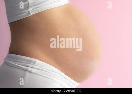 Description: Midsection of unrecognizable standing mother with very round pregnant baby belly. Side view. Pink background. Bright shot. Stock Photo