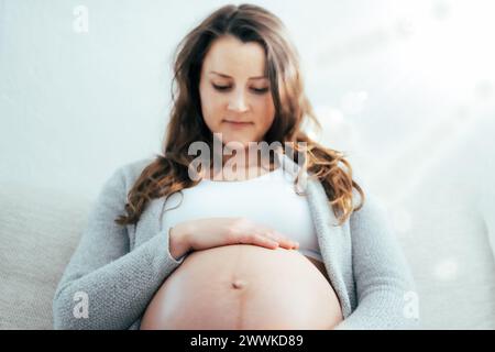 Description: Frontal view of happy woman sitting on sofa gently holding her belly in expectation of baby in last stage of pregnancy. Pregnancy third t Stock Photo