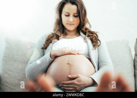 Description: Frontal view of happy woman sitting on sofa gently holding her belly in expectation of baby in last stage of pregnancy. Pregnancy third t Stock Photo