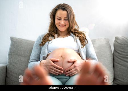 Description: Frontal view of happy smiling woman sitting on sofa gently holding her belly in expectation of having a baby in last stage of pregnancy. Stock Photo