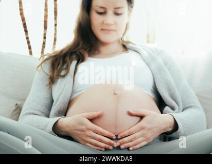 Description: Frontal view of woman sitting on sofa gently holding her belly and happily expecting her baby in last months of pregnancy. Pregnancy thir Stock Photo