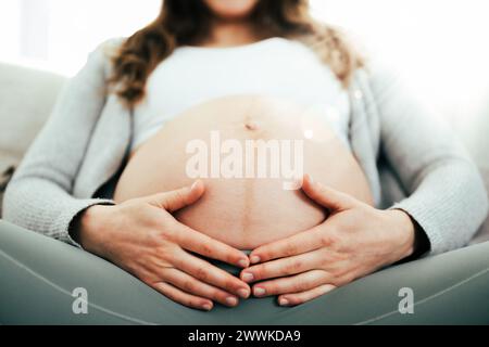 Description: Frontal view of woman sitting on sofa gently holding her belly in last months of pregnancy. Pregnancy third trimester - week 34. Frontal Stock Photo