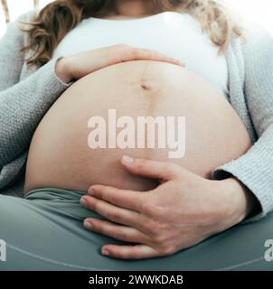 Description: Frontal view of woman sitting on sofa holding her belly in last months of pregnancy. Pregnancy third trimester - week 34. Frontal view. L Stock Photo