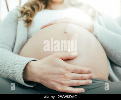 Description: Frontal view of woman sitting on sofa gently holding her belly in last months of pregnancy. Pregnancy third trimester - week 34. Frontal Stock Photo