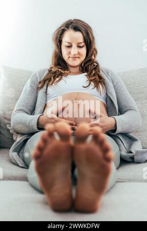 Description: Frontal view of smiling woman sitting on a sofa proudly holding her belly during the last stage of pregnancy. Pregnancy third trimester - Stock Photo