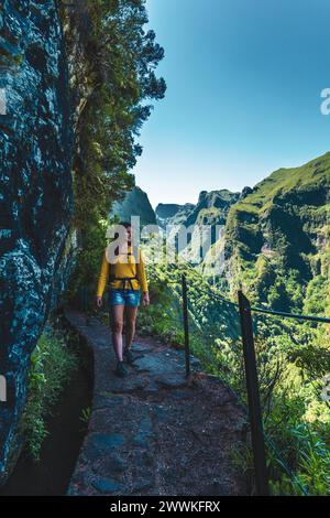 Description: Backpacker woman walking along steep cliff jungle hiking trail  next to canal through Madeiran rainforest. Levada of Caldeirão Verde, Mad Stock Photo