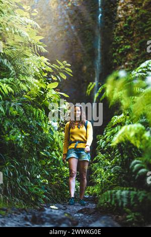 Description: Backpacker woman walking along fern overgrown trail at picturesque, overgrown waterfall in the Madeiran rainforest. Levada of Caldeirão V Stock Photo