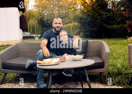 A man sitting on a black couch with his son in the backyard Stock Photo