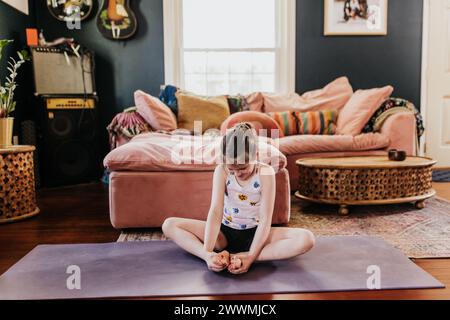 Young girl sits in butterfly pose on yoga mat in living room Stock Photo