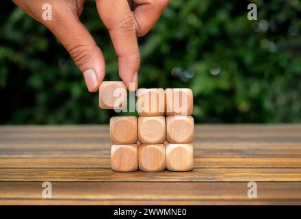 Last piece wood cube block in hand putting on wooden cube as puzzles stacking arranging completed on table and green plant background. Solution, solve Stock Photo