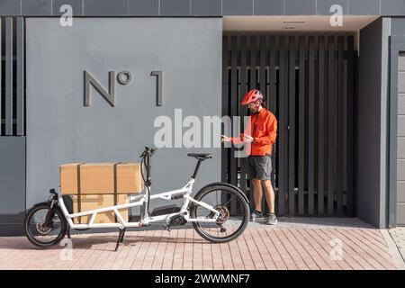 young courier with red clothing and helmet riding cargo bike arriving at the shipping destination to deliver a package to a city address, talking on t Stock Photo