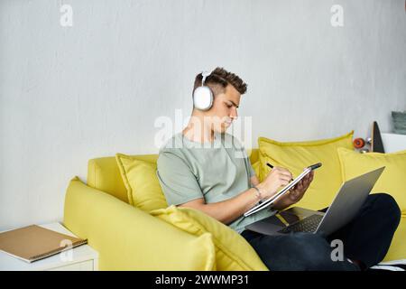 focused student with headphones and laptop in yellow couch studying and writing in note Stock Photo