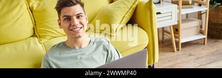 banner of student on floor near yellow couch at home sitting with laptop and smiling to camera Stock Photo