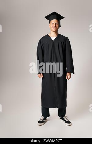 happy attractive student in graduate gown and cap standing against light background Stock Photo