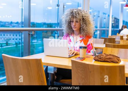 Portrait of a young Caucasian woman with curly blonde hair, working in a coffee shop with her laptop and smart phone Stock Photo