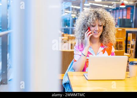 Portrait of a smiling young Caucasian woman with curly blonde hair, working in a coffee shop with her laptop and smart phone Stock Photo