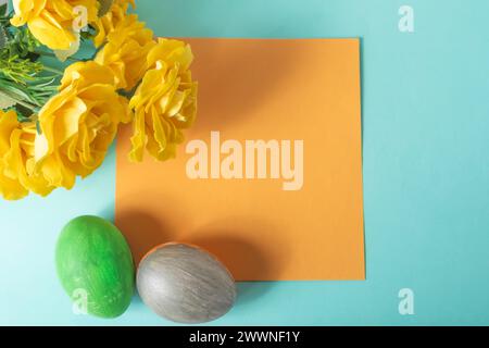 Easter eggs with yellow flowers and blank paper card on blue background with copy space. Happy Easter. Stock Photo