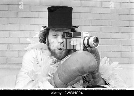 In the 1970s. A man is filming with a amateur film camera. The film was then developed and shown on a film projector on a fold up screen at home. Ricky Bruch (1946-2011) Swedish track and field athlete. He was known for being somewhat of a showman pictured here wearing boxing gloves, a feather boa and a cylinder hat.  1975 Stock Photo
