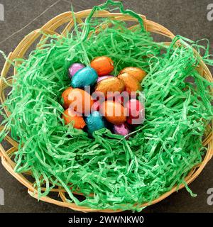 Close up on Easter Nest standing on black table, with colorful shiny chocolate easter eggs, and green grass imitation. Stock Photo