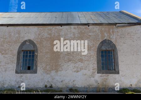 Side of an old and abandoned church building with corrugated metal roof and two gothic style windows. White paint is peeling off  the walls. Stock Photo