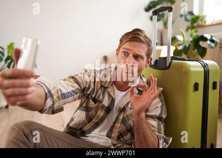 Excited young man shows his suitcase, takes selfie with okay sign on mobile phone, poses with luggage, goes on holiday Stock Photo