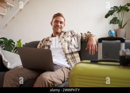 Portrait of happy young man, tourist booking his holiday with travel agency, looking at laptop, packed suitcase to go on vacation Stock Photo
