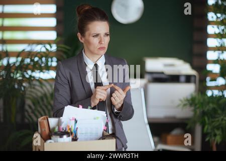 New job. pensive modern woman worker in modern green office in grey business suit with personal belongings in cardboard box. Stock Photo